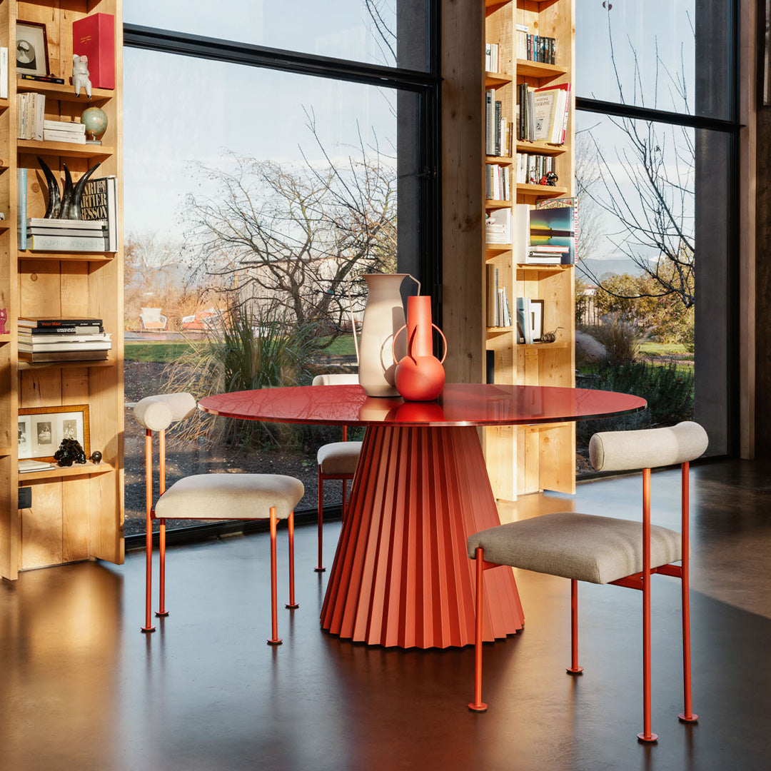 Modern orange round dining table with pleated base, complemented by white upholstered chairs and warm wooden shelves.