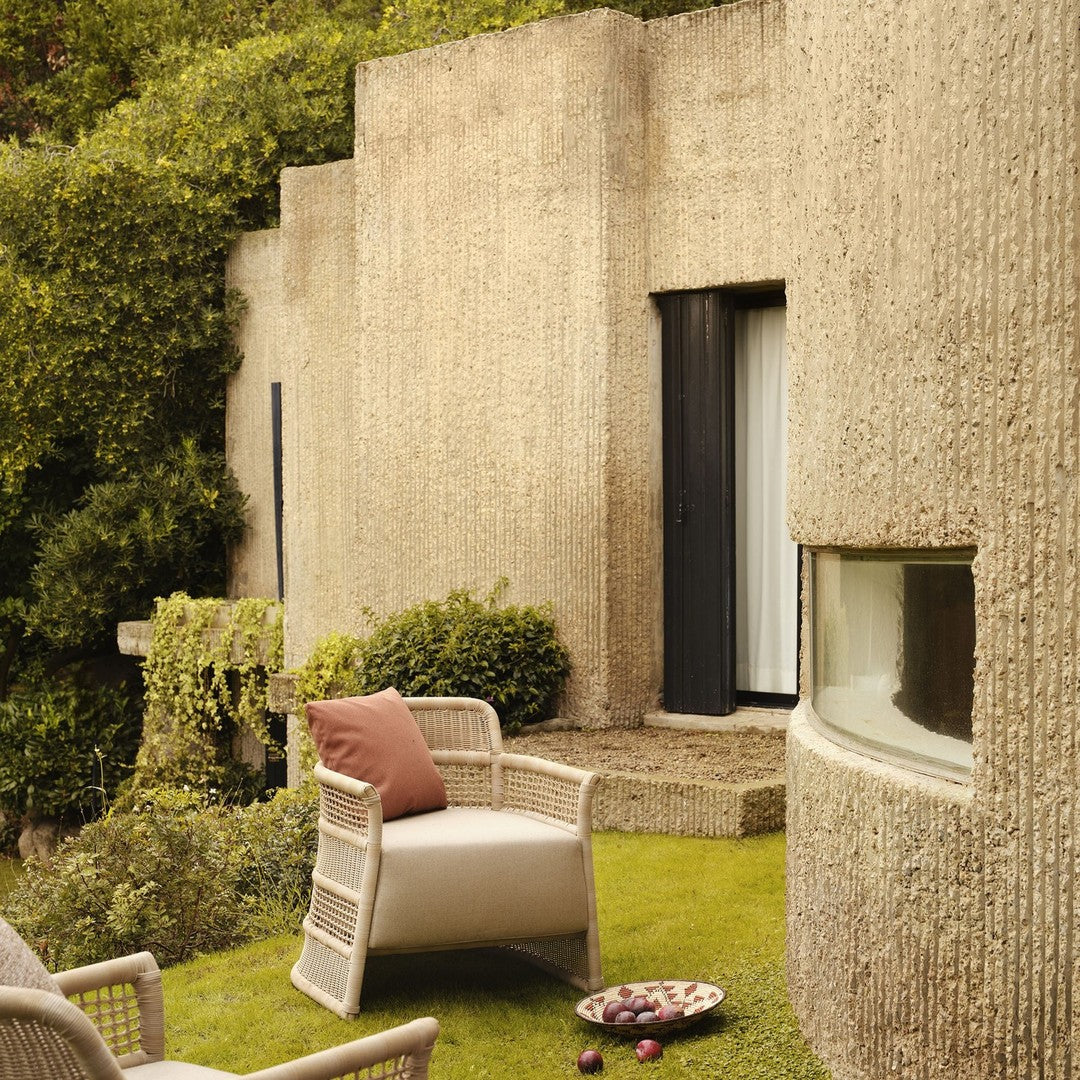 "Light gray textured patio chair with soft pink cushion, surrounded by lush green landscaping and natural stone walls."
