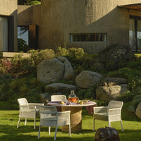 Modern outdoor dining set featuring white woven chairs and a round wooden table, set against a lush green landscape.