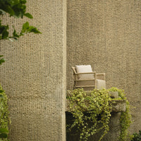 Beige wicker armchair with cream cushion, set against textured gray walls and lush green vines. Ideal for outdoor spaces.