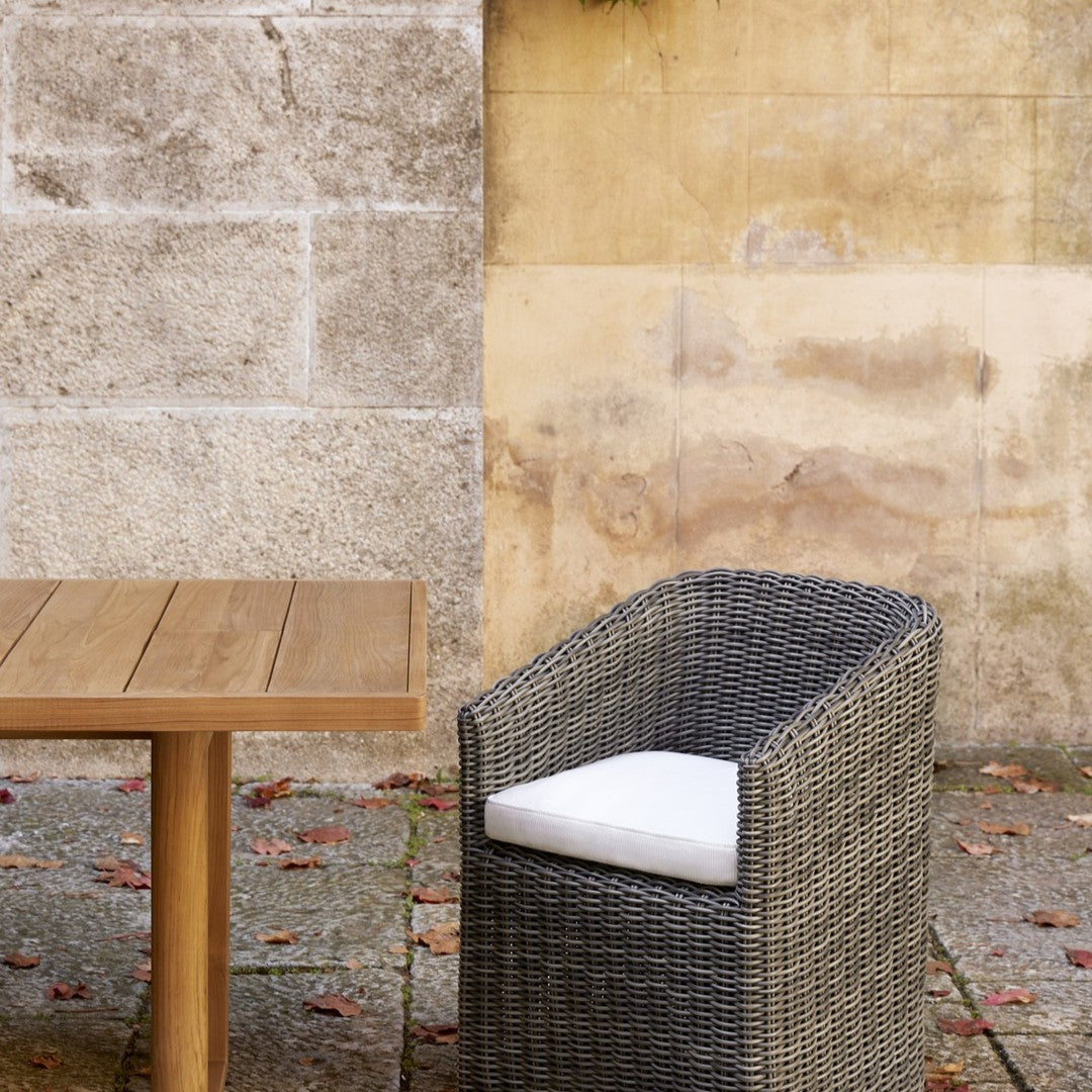 Gray wicker chair with white cushion beside a wooden table against a neutral stone wall.