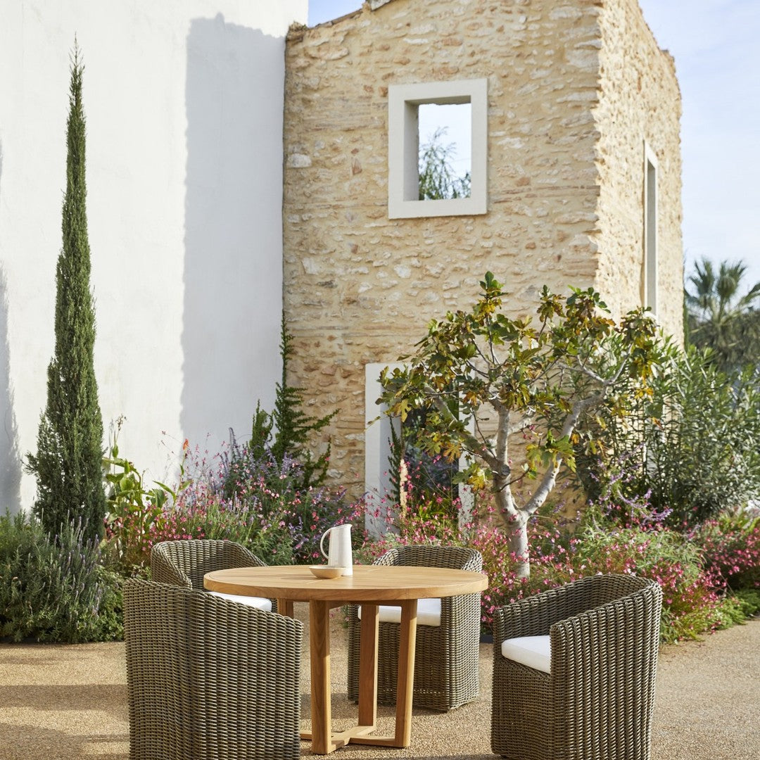 Natural stone backdrop with greenery; round wooden table surrounded by textured gray wicker chairs; vibrant floral accents.