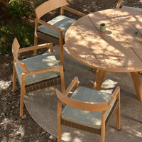 Natural wood chairs with light grey cushions and a round wood table, set on a textured outdoor rug.