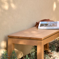 Light wood bench with smooth planks, complemented by earthy beige wall and greenery. Simple, modern design.