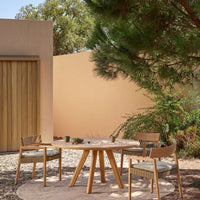Natural wood table and chairs with green cushions, set against a soft peach wall and lush greenery. Perfect for outdoor dining.