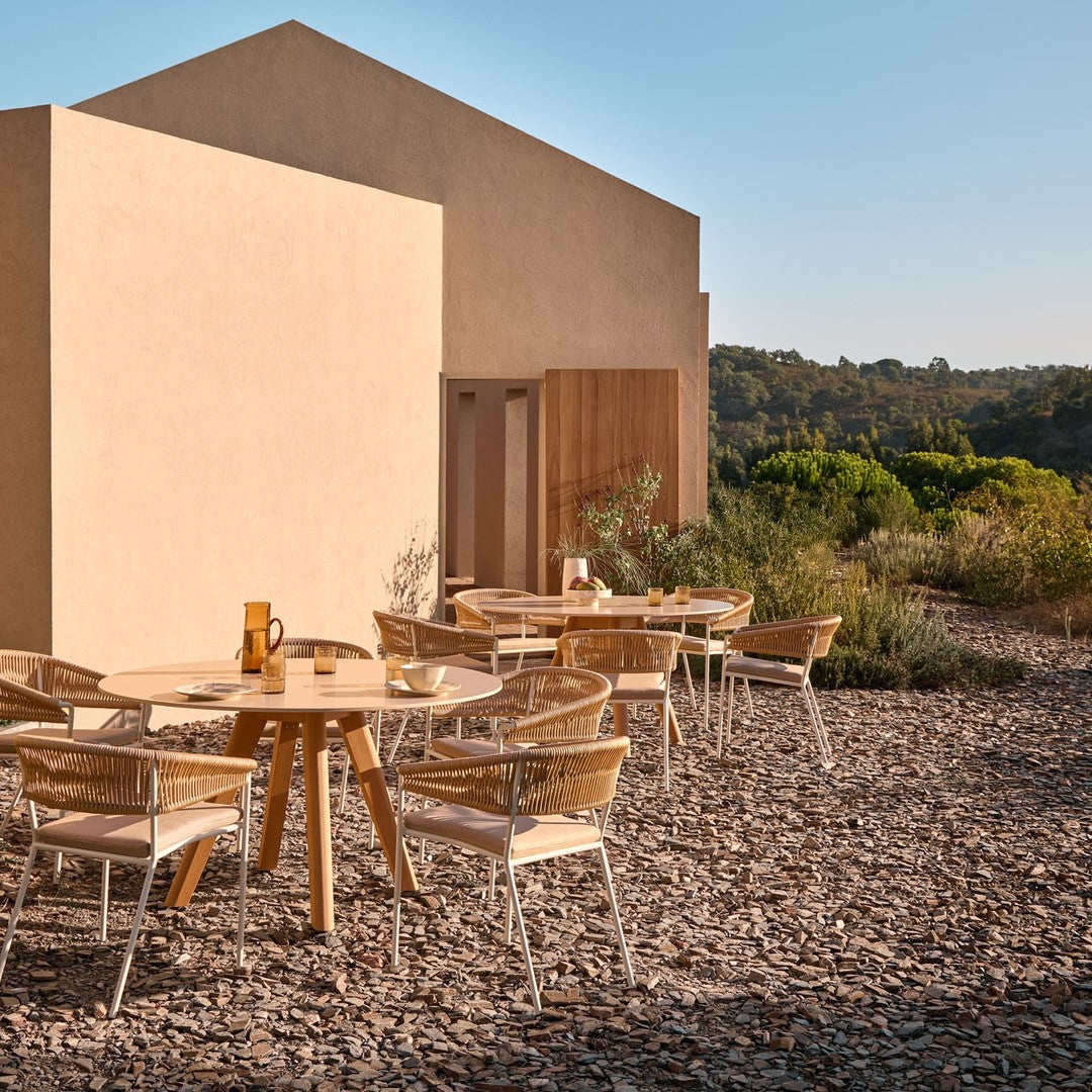 Wooden table and chairs with neutral tones, set against earthy stone textures and a minimalist building backdrop.