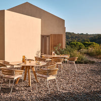 Wooden table and chairs with neutral tones, set against earthy stone textures and a minimalist building backdrop.