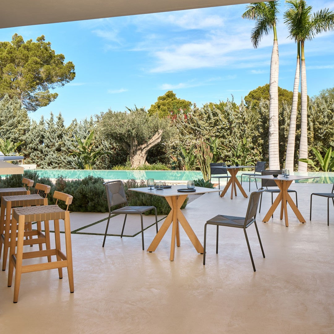 "Modern outdoor furniture set featuring gray chairs, wooden tables, and lush green foliage against a blue sky backdrop."
