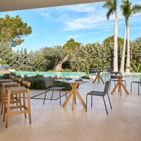 "Modern outdoor furniture set featuring gray chairs, wooden tables, and lush green foliage against a blue sky backdrop."