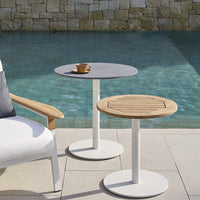 "Modern round tables with grey and wood finishes, featuring white metal bases, near a serene pool backdrop."