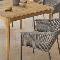 Natural wood dining table with a light finish and a grey woven chair, perfect for modern outdoor settings.