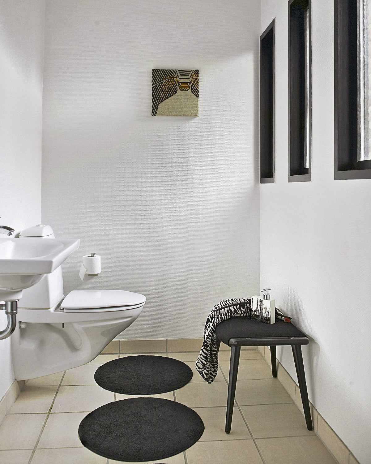 Black round bathroom rugs on a light tan tiled floor, surrounded by white walls and dark window frames.