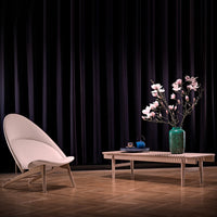 Light beige modern chair and light wooden coffee table against a dark purple curtain backdrop, accented with pink floral arrangement.