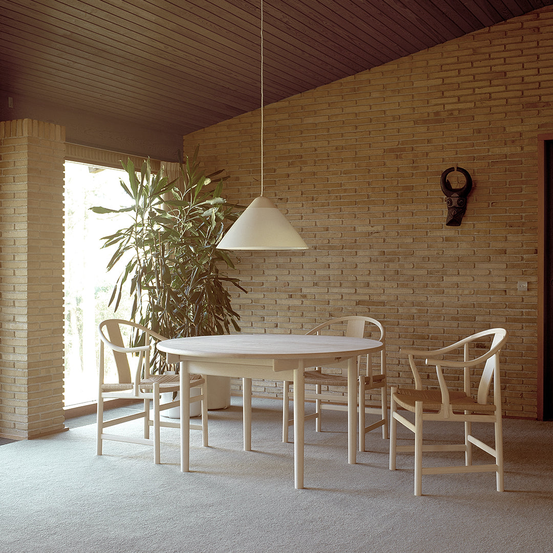 Light wood dining table and chairs with a simple, neutral design; warm brick wall and soft natural lighting.