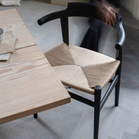 Black chair with a woven beige seat, featuring a diagonal pattern; natural wood table in a neutral-toned setting.