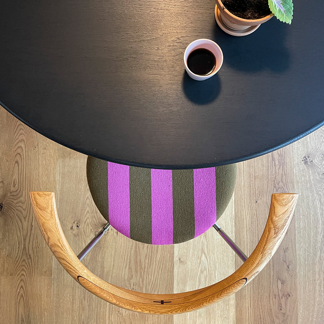 Top view of a black round table with a green striped stool in purple and green, featuring a wooden chair arm and plants.