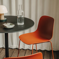 Orange chair with a smooth finish, black round table, and neutral curtains in a minimalist setting.