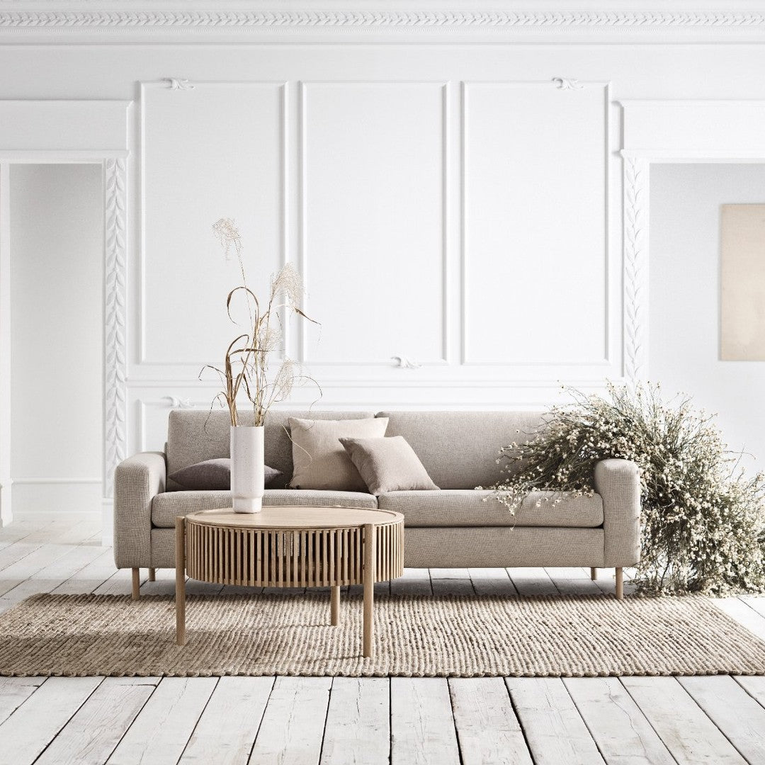 Light gray sofa with subtle texture, neutral cushions, and a round wooden coffee table on a woven rug against white walls.