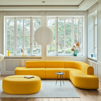 Bright yellow curved sectional sofa with coordinating round ottoman, set against a light, airy room with large windows.