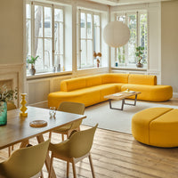 Modern living room featuring a vibrant yellow sectional sofa, round ottoman, and wooden dining table in a light interior.
