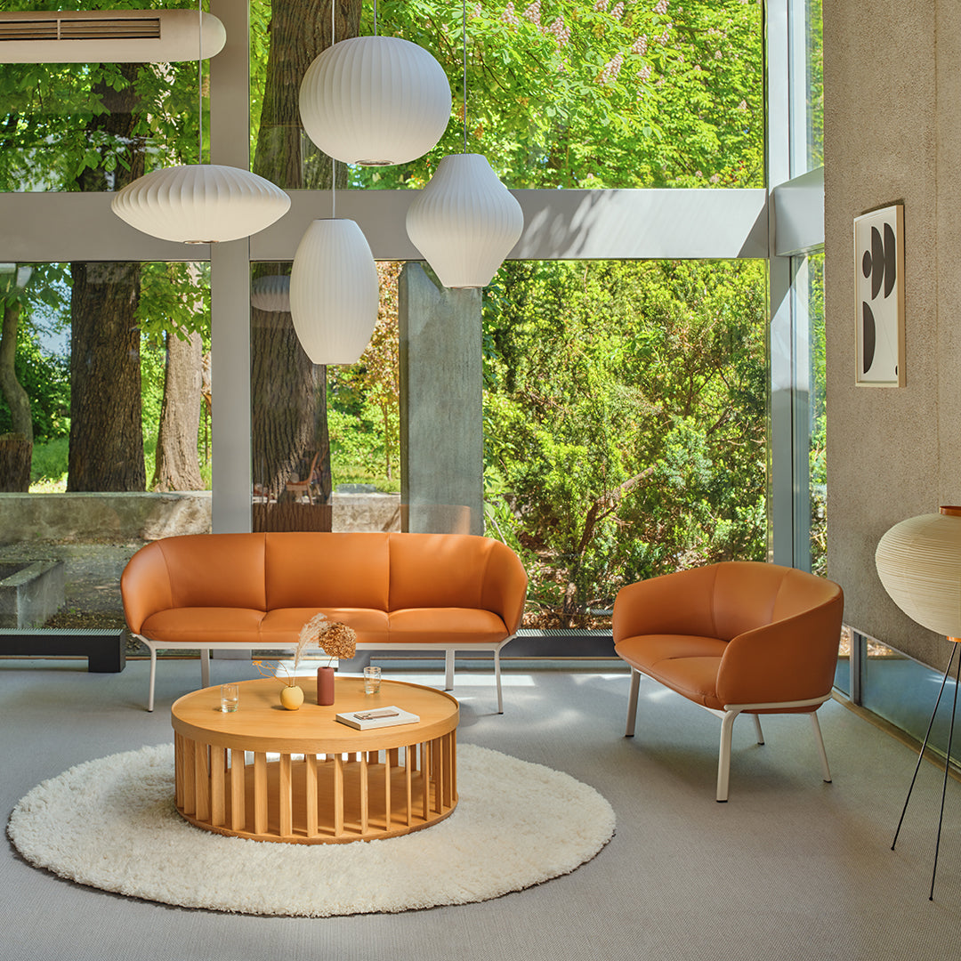 "Modern living room with orange sofa, light brown chairs, round wooden coffee table, and white pendant lights."