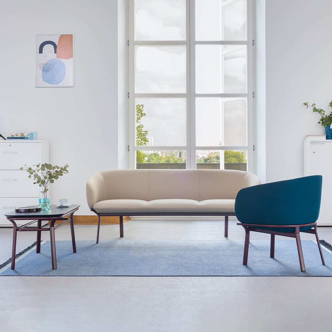 Beige sofa with teal accent chair, gray-blue rug, and minimalist decor in a bright, airy room.