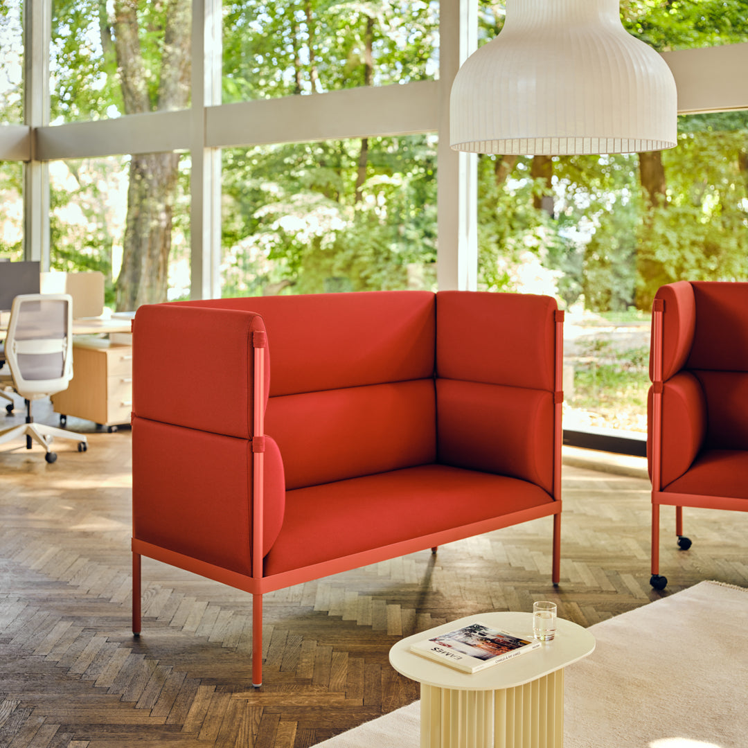 Modern red upholstered two-seater sofa with clean lines and minimalistic design in a bright, leafy setting.
