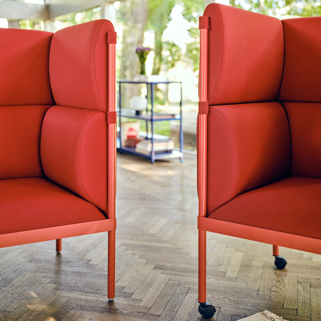 Pair of modern red upholstered chairs with sleek design, set against a wooden floor and lush greenery backdrop.