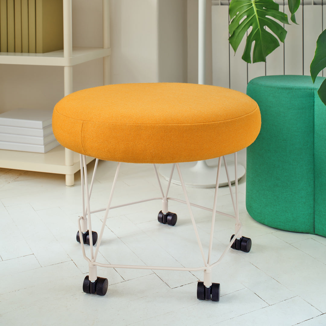 Round orange upholstered ottoman on wheels with a minimalist design and white metal wire base, complemented by a green pouf.