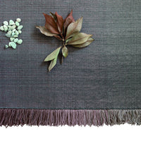 Gray woven textile with subtle gradient and fringe, complemented by green and brown botanical accents.
