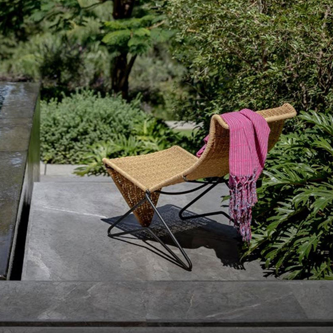 "Natural woven rattan chair with a vibrant pink fringed throw, surrounded by lush green foliage."