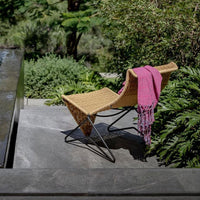 "Natural woven rattan chair with a vibrant pink fringed throw, surrounded by lush green foliage."