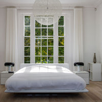 Bright white bedding on a modern bed, surrounded by light wood flooring, with green views through large windows.
