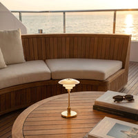 Elegant gold and white lamp on wooden table, with soft beige cushions and natural wood patterns, set against a sunset backdrop.