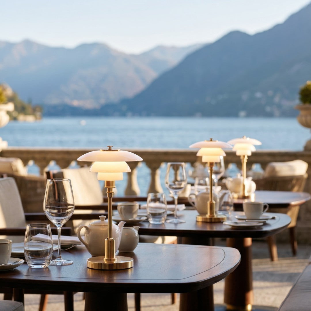Elegant white and gold table lamps contrast against the natural tones of a scenic outdoor dining setup by the water.