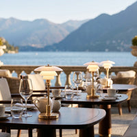 Elegant white and gold table lamps contrast against the natural tones of a scenic outdoor dining setup by the water.