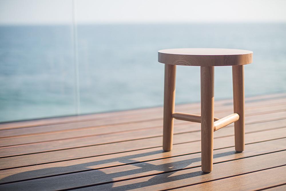 Natural wood stool with a smooth finish, featuring a simple round seat and sturdy legs against a serene ocean backdrop.