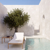 "Relaxing outdoor scene with a white lounge chair, wooden table, olive tree, and textured beige walls near a clear pool."
