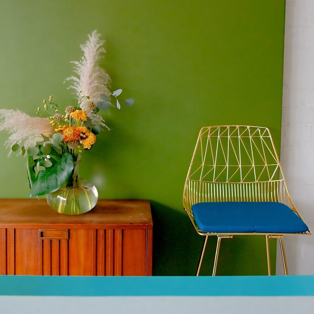 Gold wire chair with a blue seat against a vibrant green wall; features a wooden cabinet and colorful floral arrangement.