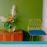 Gold wire chair with a blue seat against a vibrant green wall; features a wooden cabinet and colorful floral arrangement.