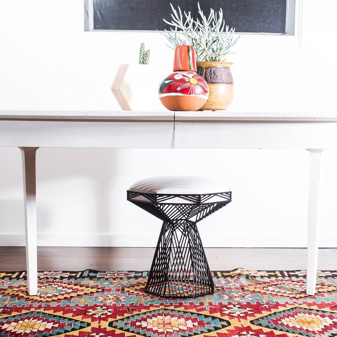 Stylish black wire stool on a colorful, patterned rug with red, yellow, and green hues. Minimalist white table and decor.