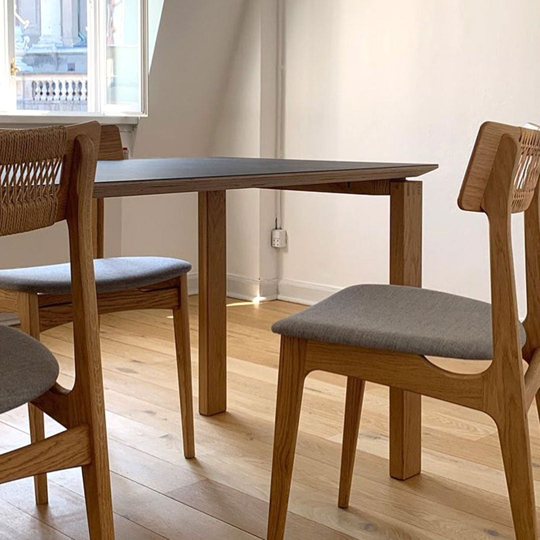 Light wood dining table with textured beige chair upholstery and woven backrest, set against a bright, minimalistic backdrop.