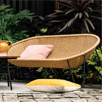 Stylish rattan loveseat featuring a natural beige color, pink accent cushion, and vibrant yellow pillow, set in greenery.