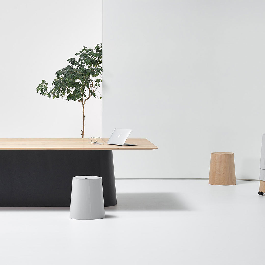 Modern office setup with a light wood desk, black base, gray and natural wood stools, and a minimalist white backdrop.