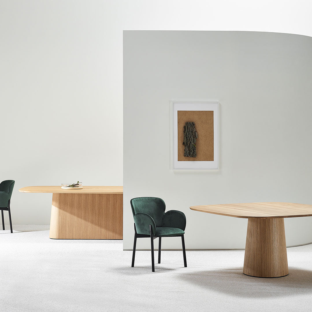 Modern interior featuring green velvet chair, light wood tables, white walls, and a textured brown artwork.