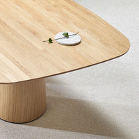 Natural wood coffee table in light oak finish with oval-shaped top and textured beige rug underneath.