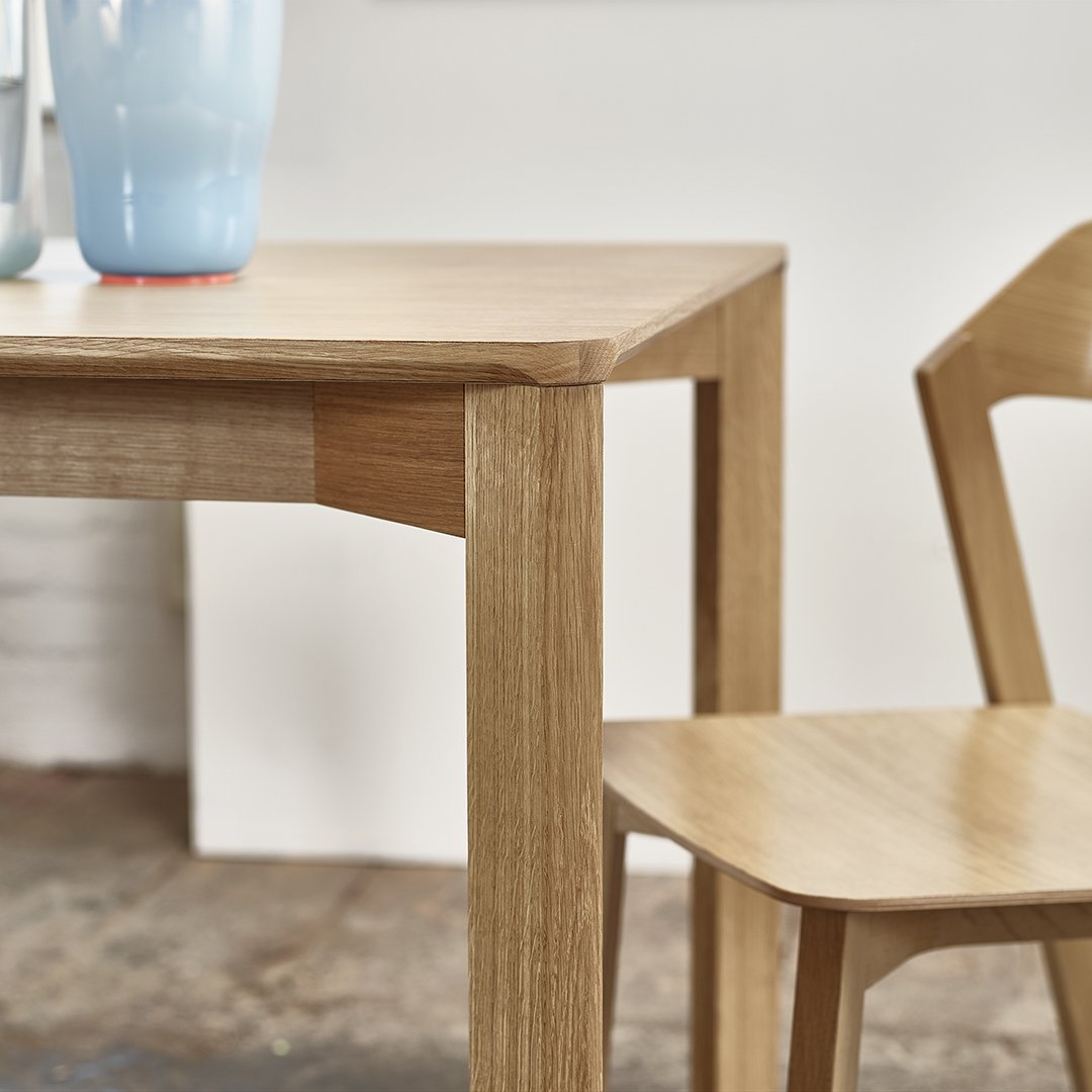 Light oak wooden table with a simple design, paired with a matching light oak chair, set against a neutral backdrop.