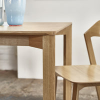 Light oak dining table and chair with natural wood grain, accented by a soft blue vase. Modern, minimalist design.