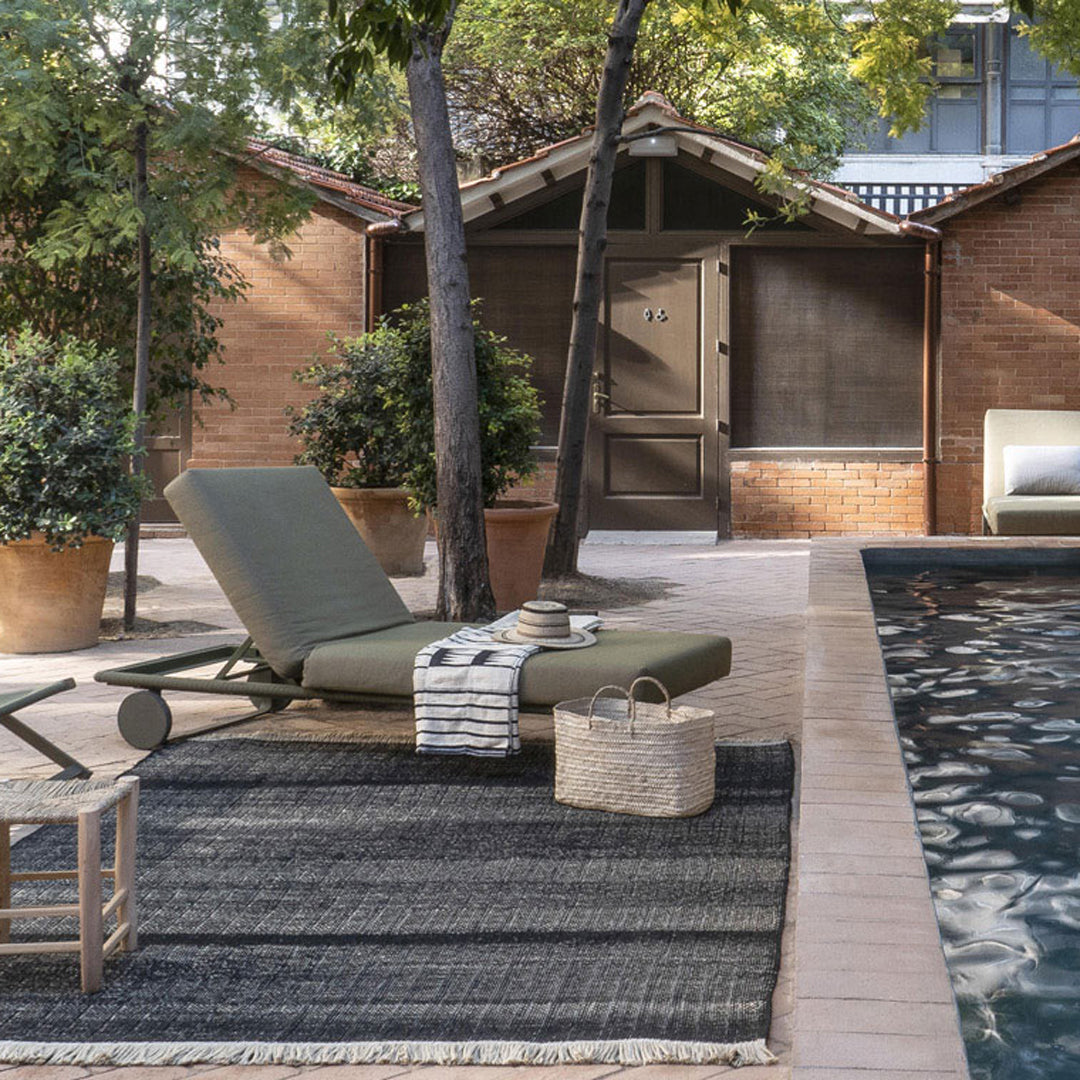 Gray and green lounge chair on a textured black rug, surrounded by greenery and earthy tones in a serene outdoor setting.