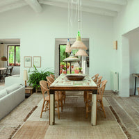 Modern dining space featuring beige woven rugs, green plants, and colorful pendant lights against a neutral background.
